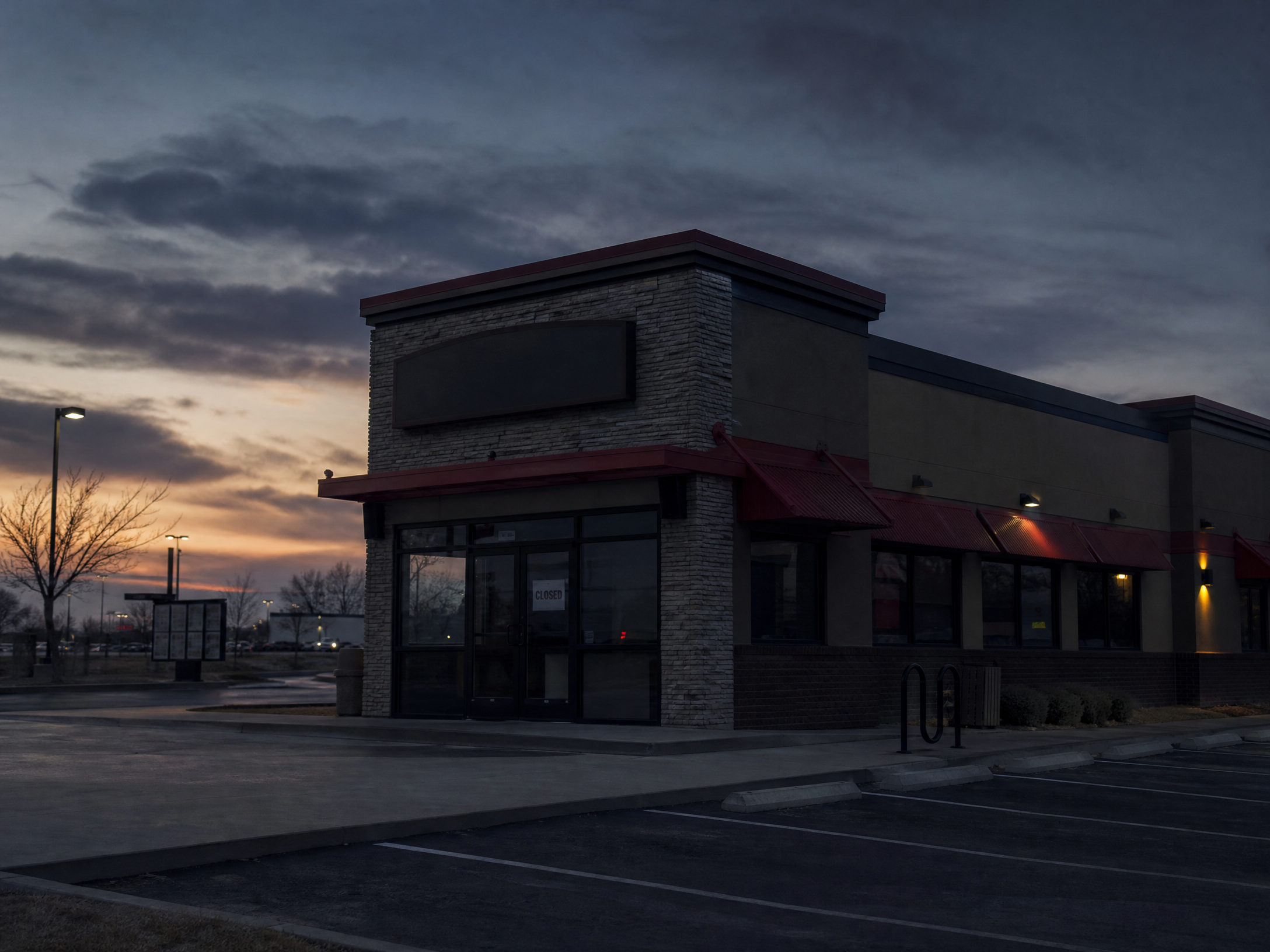A closed fast food restaurant with a darkened storefront, 'Closed' sign on the door, empty parking lot at dusk. The building looks like a typical American fast food chain exterior. Moody atmospheric lighting, street photography style, no text overlay